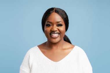 Genç siyahi kadın neşeyle gülümsüyor, mutluluğu ve pozitifliği mavi bir stüdyoda ifade ediyor.