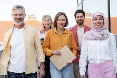 Çok uluslu gülümseyen iş adamları ve iş kadınlarından oluşan çeşitli bir grup dışarıda poz veren kameralara bakıyor. Takım çalışması konsepti