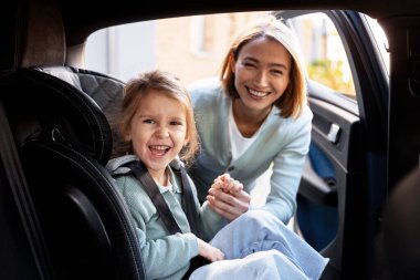 Anne ve kızı gülümserken çocuk bir araba koltuğuna oturmuş yolculuk ederken.
