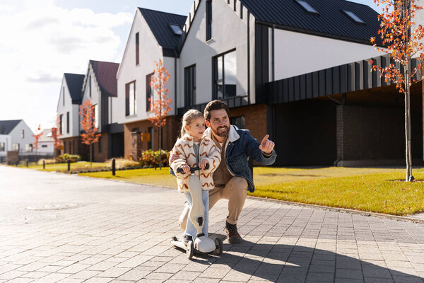 Father teaching young daughter to ride a scooter outdoors on a paved street