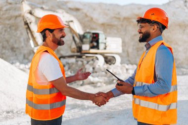 Turuncu şapka ve yelek giyen profesyonel inşaat işçileri taş ocağında el sıkışıyor. Ortaklık, takım çalışması konsepti