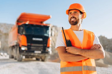 Turuncu güvenlik yeleği giymiş yakışıklı inşaat ustabaşı dışarıda kollarını kavuşturmuş, endüstriyel alanda süreci izliyor. Ağır makineler kavramı