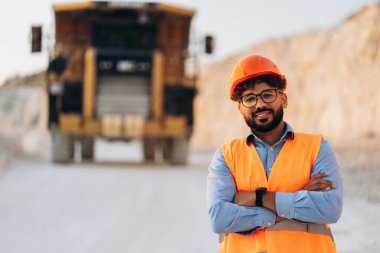 Orta Doğulu gülümseyen, turuncu yelek giyen ve kolları çapraz olan kasklı bir adam taş ocağında kameraya bakıyor. Ağır sanayi konsepti