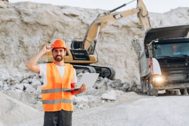 Koruyucu ekipman giyen erkek site yöneticisi bir taş ocağında dizüstü bilgisayar kullanıyor, kameraya bakıyor, kopyalama alanı. Çevrimiçi teknoloji kavramı