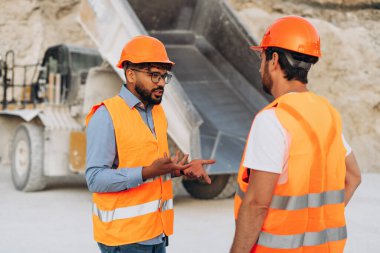 Ağır çöp kamyonunun yanındaki taş ocağında turuncu güvenlik yeleği ve kask giyen iki inşaat işçisi. Takım çalışması konsepti
