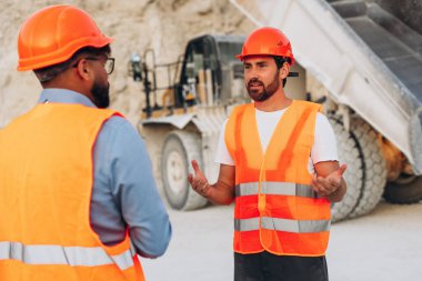 Ağır çöp kamyonunun yanındaki taş ocağında turuncu güvenlik yeleği ve kask giyen iki inşaat işçisi. Takım çalışması konsepti
