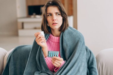 Kendini hasta hisseden genç bir kadın, elinde mendil ve ilaç, kanepeye sarılmış bir şekilde.