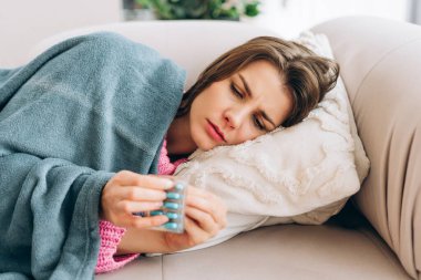 Kanepede uzanmış bir kadın hastalıkla boğuşuyor ve su toplamış bir paketten ilaç seçiyor.