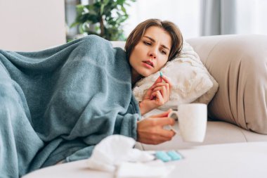 Bir kadın battaniyenin altında kanepeye uzanıyor, bir hap alıyor ve sıcak bir içecek içiyor.