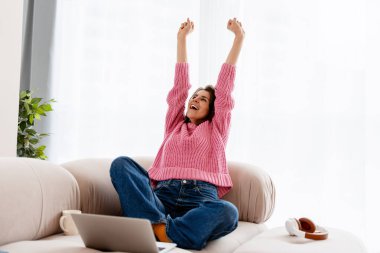 Başarı kutlayan bir kadın, dizüstü bilgisayarla koltukta çalışıyor.