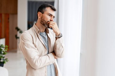 Pencerenin yanında duran bir adam, bir sorun üzerinde kafa yoruyor, elleri çenesinde.