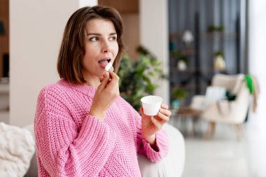 Oturma odasındaki bardaktan yoğurt yiyen bir kadın sağlıklı bir atıştırmalıkla tatmin olur.