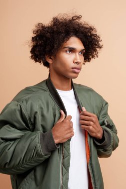 Siyah kıvırcık saçlı ve burnu piercing 'li genç adam yeşil bir bombacı ceketi giyiyor, bej bir arka plana yan bakıyor.