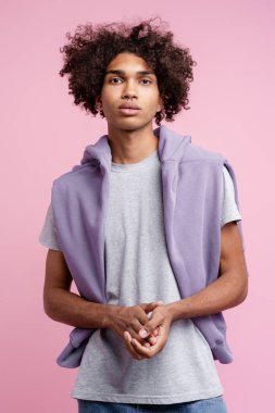 Stüdyoda kıvırcık saçlı, sıradan tişörtlü, omuzlarında kapüşonlu, burun halkası, pembe arka planı olan genç bir adam.