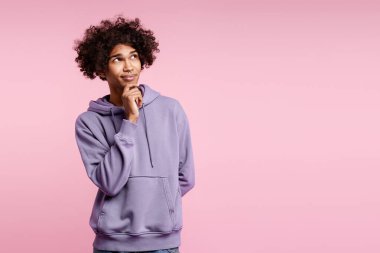 Kıvırcık saçlı genç bir adam elini çenesine koyuyor, pembe bir arka plana bakıyor.