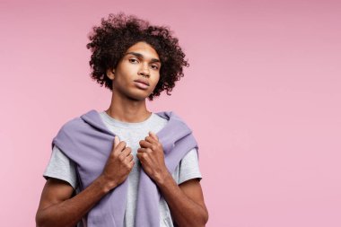 Kıvırcık saçlı ve burun halkalı genç bir adam omuzlarının üzerine pembe bir süveter giyiyor.