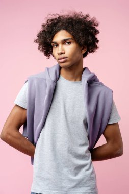 Kıvırcık saçlı, kendine güvenen bir genç. Gri bir tişört giyiyor ve omuzlarının üzerine de mor bir kapüşonlu kazak giyiyor.