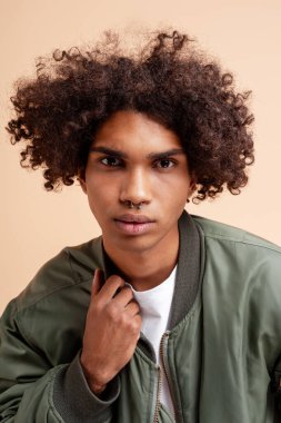 Kıvırcık saçlı, burnu piercing takan, bombardıman ceketli, düz tişörtlü genç bir adam.