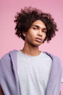 Kıvırcık saçlı, burnu piercing 'li, gri tişörtlü genç bir adam stüdyoda güven içinde poz veriyor.