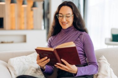 Gülümseyen, gözlüklü, elinde kitap okuyan mutlu esmer kadın evdeki rahat koltukta oturuyor. Eğitim kavramı