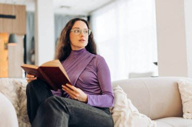 Güzel, mutlu, gözlüklü, kitap okuyan esmer kadın evdeki rahat koltukta oturuyor. Eğitim kavramı