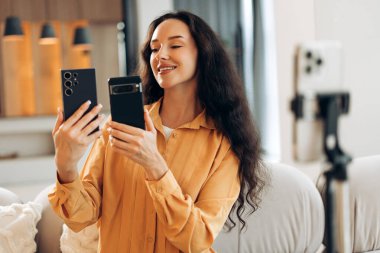 Neşeli, gülümseyen, uzun saçlı, elinde cep telefonu olan beyaz bir kadın akıllı telefonun fotoğrafını çekiyor, blogger videosu kaydediyor. Çevrimiçi teknoloji kavramı