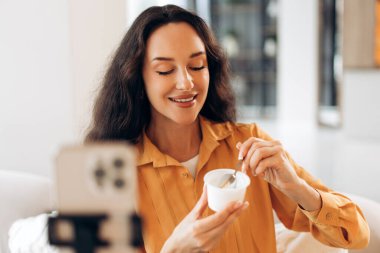 Pozitif, gülümseyen bir kadın elinde yoğurt ve kaşıkla beyaz bardak tutuyor, video kaydediyor, cep telefonu kullanıyor, beslenme uzmanı konuşuyor. Dengeli diyet kavramı