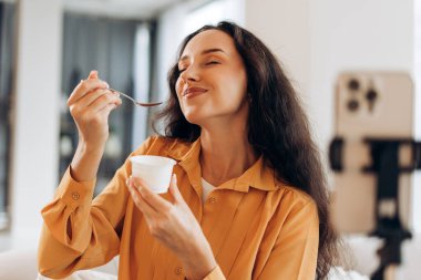 Uzun siyah saçlı neşeli bir kadın lezzetli bir tatlı yiyor, elinde beyaz bardak ve kaşık tutuyor, blogger evinde video kaydediyor.