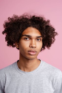 Yakışıklı, şık Afrikalı Amerikalı genç adam. Burnuna piercing taktırmış ve kıvırcık saç stiliyle kameraya bakıyor. Pembe arka planda izole edilmiş bir model.