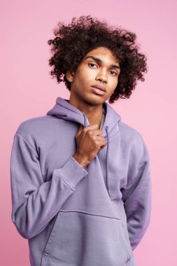 Yakışıklı, kıvırcık kıvırcık saçlı, şık mor kapüşonlu bir adam pembe arka planda yalnız poz veren kameraya bakıyor. Moda konsepti