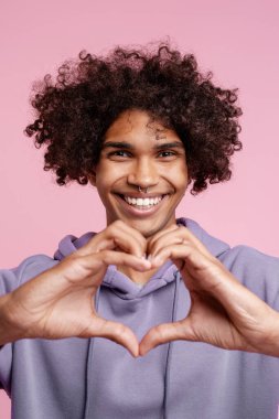 Yakışıklı, gülümseyen, burnunu delen, şık kapüşonlu, şömineyi elleriyle gösteren pembe arka planda yalnız poz veren Latin bir genç. Aşk, reklam konsepti