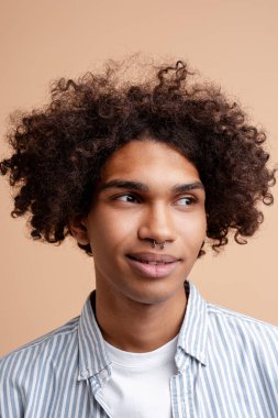 Gülümseyen, otantik İspanyol kıvırcık saçların portresi. Burnu delinmiş, bej arka planda izole bir şekilde uzağa bakıyor. Reklam kavramı