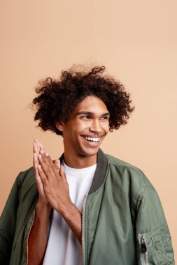 Mutlu, yakışıklı, otantik, burnunu delen, şık yeşil ceket giyen, diş tebessümü ile bej arka planda yalnız poz veren genç bir adam. Alışveriş konsepti