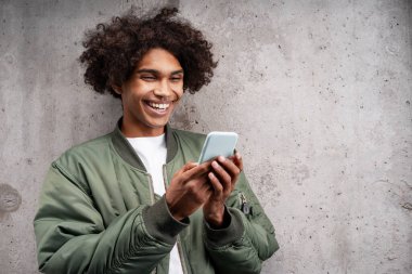 Cep telefonunda piercing 'i olan mutlu Afro-Amerikalı bir adamın portresi. Gri arka planda izole edilmiş mobil uygulama metin kopyalama uzayı kullanıyor. Çevrimiçi alışveriş kavramı