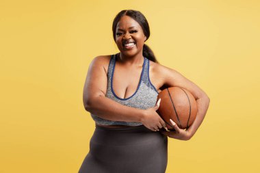Gülümseyen ve büyük boy bir Afro-Amerikan modeli şık bir bluz giyerek basketbol topunu tutuyor kameraya bakıyor, sarı arka planda izole bir şekilde poz veriyor. Spor konsepti