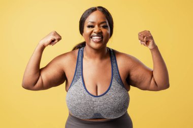 Heyecanlı gülümseyen Afro-Amerikan kadın şık giyinen güçlü elleri ve sarı arka planda izole edilmiş pazıları olan pozitif kadın. Spor konsepti