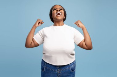 Mutlu, heyecanlı, iri yarı, sıradan kıyafetler giyen Afro-Amerikan kadın mavi arka planda izole edilmiş paralara bakıyor. Zafer konsepti