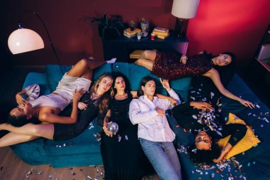 Çeşitli genç yetişkinler bir kanepede uzanıp neşeli bir kutlamanın ardından dinleniyorlar.