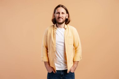 Uzun saçlı ve sakallı bir adam gülümsüyor, arka planda elleri ceplerinde duruyor.