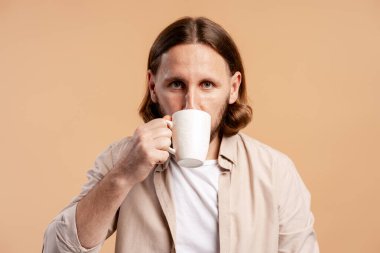 Beyaz bardaktan kahve içen bir adam, sıradan bir ev ortamında kameraya bakıyor.