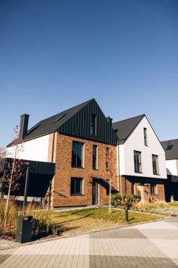 Modern banliyö evlerinde tuğla ve beyaz cepheli modern mimari tasarım, karanlık çatılar ve berrak mavi gökyüzü altında manikürlü peyzaj sergileniyor.
