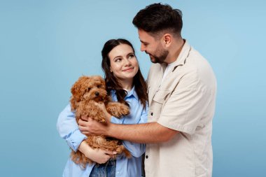 Genç çift kucaklaşıyor, yeni köpeklerini kucaklıyorlar, mavi bir arka planda mutluluk ve şefkat gösteriyorlar.