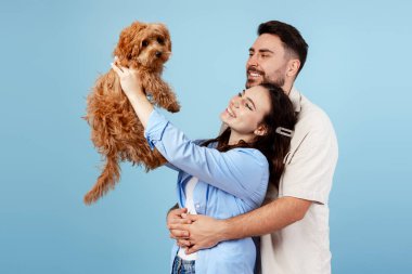 Sarılan genç çift, sevimli bir köpeği kaldıran kadın, mavi arka planda mutluluk ve aile bağı gösteren.