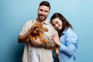 Genç bir çift gülümsüyor, dekoratif ışıkları olan küçük bir köpeği kucaklıyor, neşeyi ve birlikteliği simgeliyor.