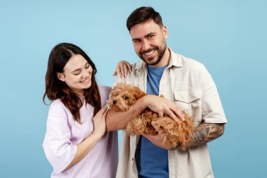 Gülümseyen adam ve kadın mavi arka planda küçük kahverengi bir köpek yavrusu tutuyorlar, evcil hayvan sevgisini ifade ediyorlar.