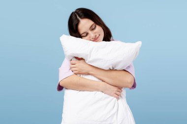 Genç bir kadın yorgun ve rahat hissediyor, günlük pijamalarıyla yastığına sarılıyor.