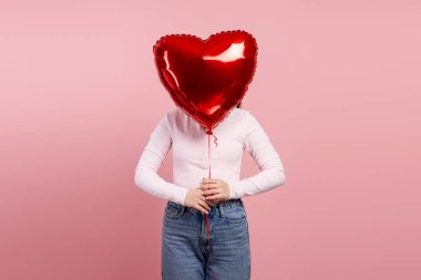 Pembe arka planda parlak kırmızı bir kalp balonuyla yüzünü saklayan genç bir kadın aşkı ifade ediyor.