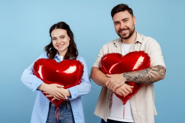 Genç çift gülümsüyor, kırmızı kalp balonlarını kucaklıyor, romantizmi ve özel günleri kutluyor.