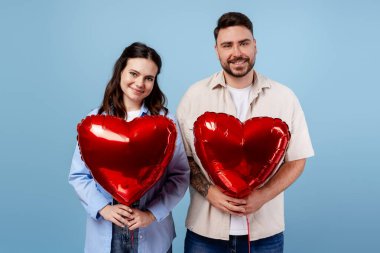 Genç adam ve kadın gülümsüyor, ellerinde kırmızı kalp şeklinde balonlar var mavi arka planda.