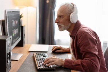 Beyaz kulaklıklı olgun bir adam masaüstü bilgisayarıyla çalışıyor ya da dinleniyor